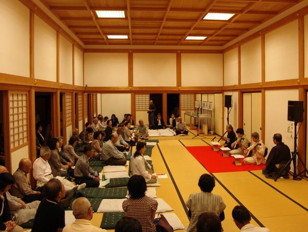 着物を着た3名の女性が三味線を弾いており、その様子を真剣に鑑賞している方々の写真